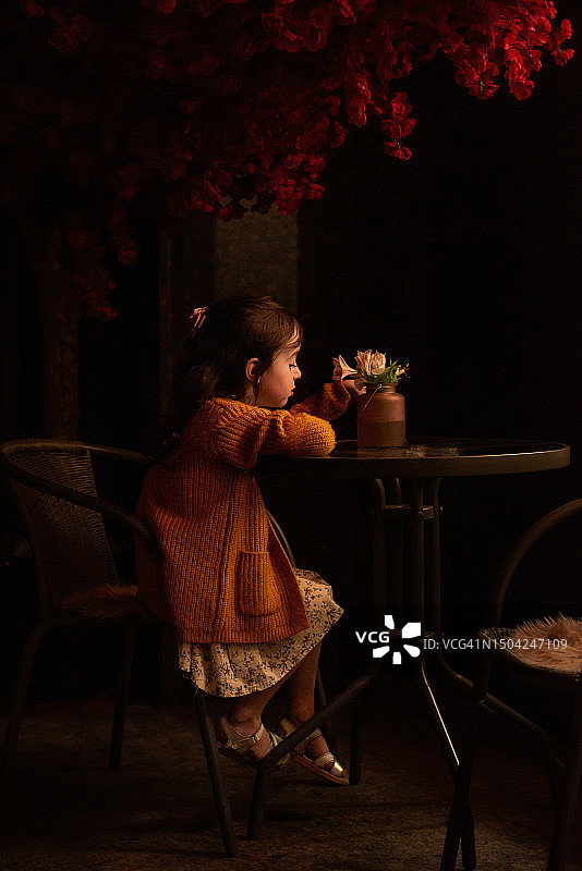 秋天傍晚，穿着芥末色针织开衫的小女孩坐在咖啡馆的桌子旁，背景是红色的树图片素材