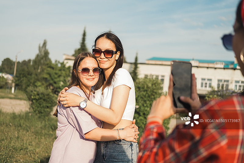 三个年轻的白人女孩一起享受户外假期。右边的黑发女孩用手机给她的朋友们拍照。生活方式概念。图片素材