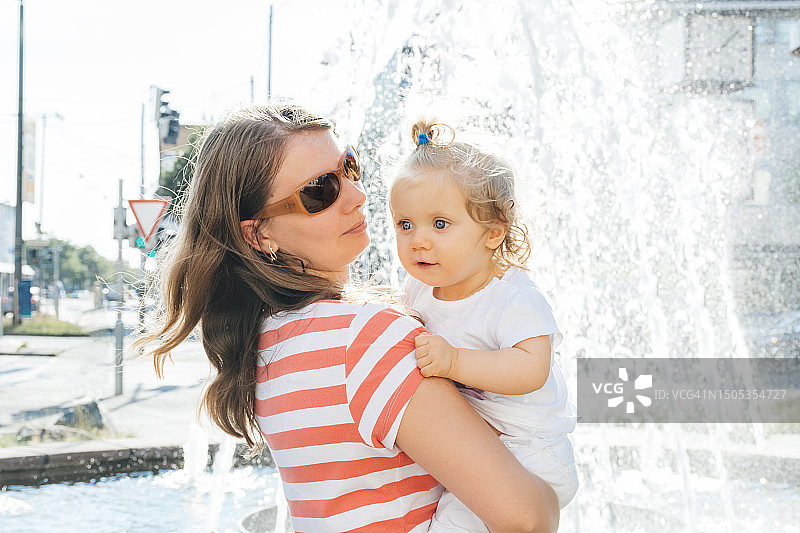 快乐的母亲抱着微笑的女儿在喷泉旁图片素材