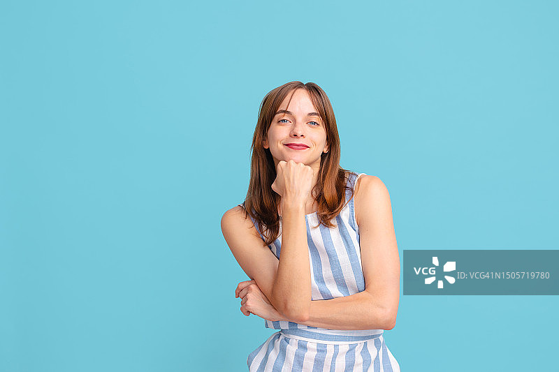 身穿条纹连衣裙的年轻女人对着镜头微笑，用手托着下巴，站在蓝色的工作室背景前图片素材