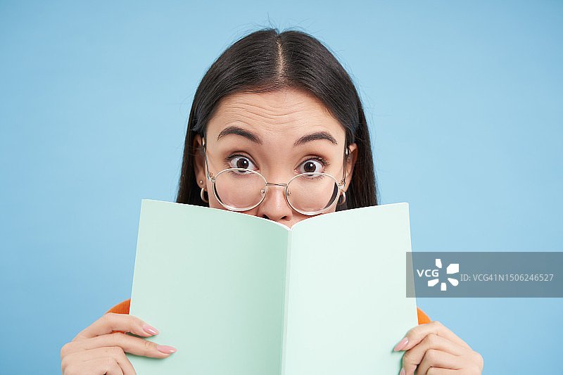 用书遮住嘴巴的年轻女子肖像，蓝色背景图片素材