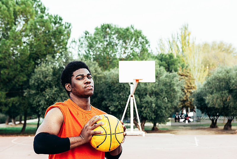 黑人男孩在城市篮球场上抱着篮球的肖像，穿着橙色T恤图片素材