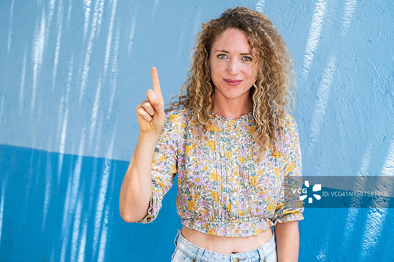 年轻金发卷发女人微笑，看着镜头，比“第一”手势图片素材