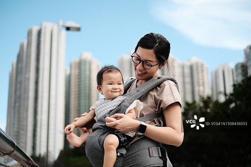 充满爱意的年轻亚洲母亲在城市中抱着婴儿女儿，背景是高层建筑图片素材