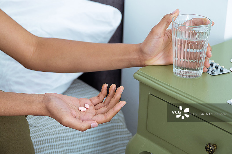 一位年轻的深色皮肤女士手握一杯水，在床上吃药，在家里的卧室里感觉不舒服。一位服用药物或止痛药的妇女，患有头痛、偏头痛、抑郁症。医疗。宿醉综合征。图片素材