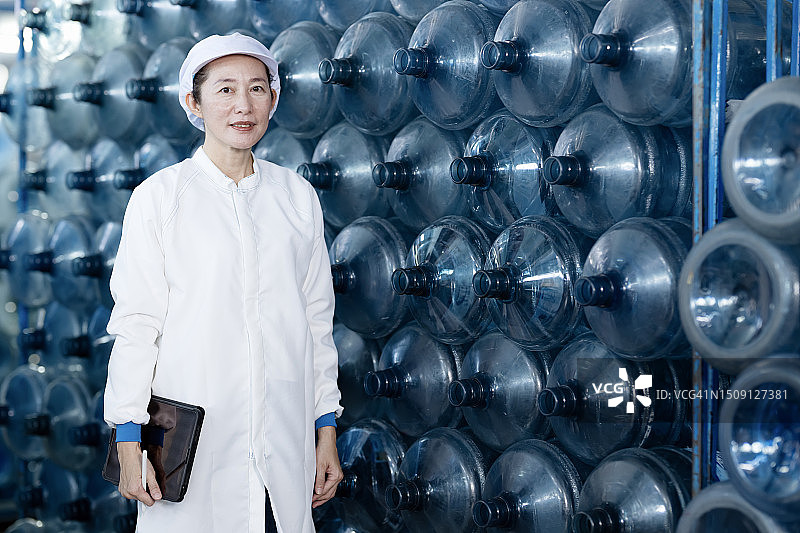 水灌装行业中可重复使用水桶的安全和处理清洁过程。一位日本女性饮用水厂经理站在堆叠的可重复使用水桶旁，测量清洁过程中的质量。图片素材