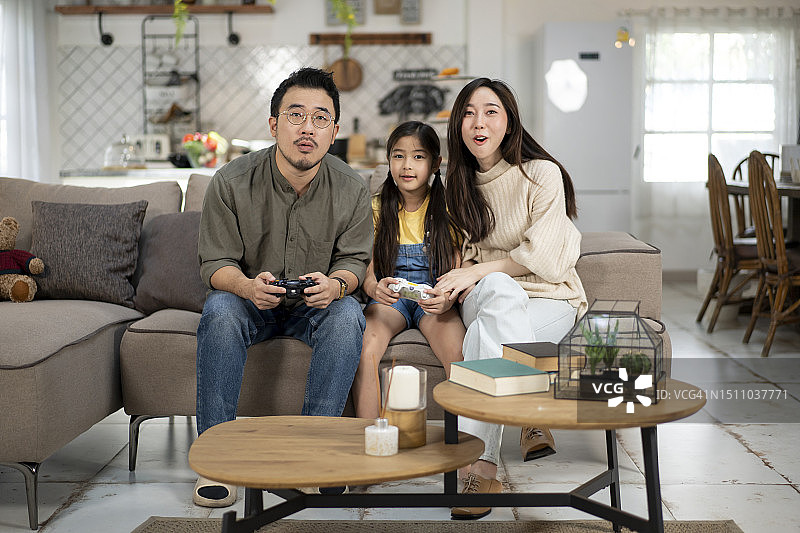 快乐的年轻亚洲家庭和女儿在客厅的沙发上玩电子游戏，共度美好时光图片素材
