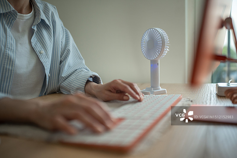 女人在电脑键盘上打字，便携式风扇在桌旁降温图片素材