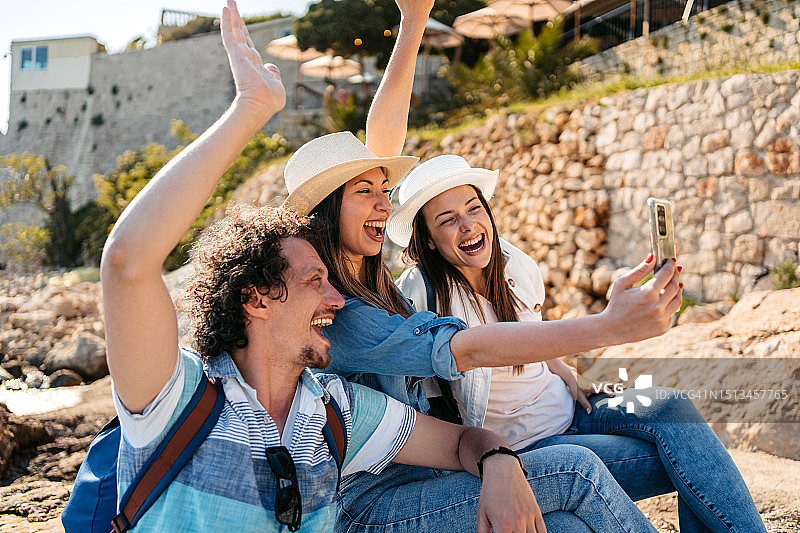 三个年轻的朋友坐在杜布罗夫尼克海湾的岩石上自拍图片素材