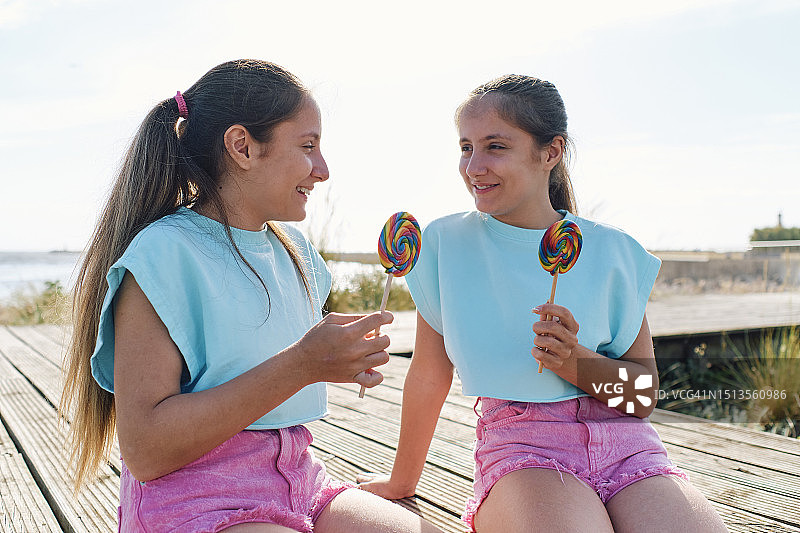 快乐的女孩们拿着棒棒糖坐在木板路上图片素材