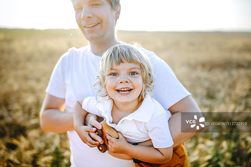 温馨的父爱：父亲在宁静的麦田上拥抱幼儿儿子，一起玩木制玩具图片素材