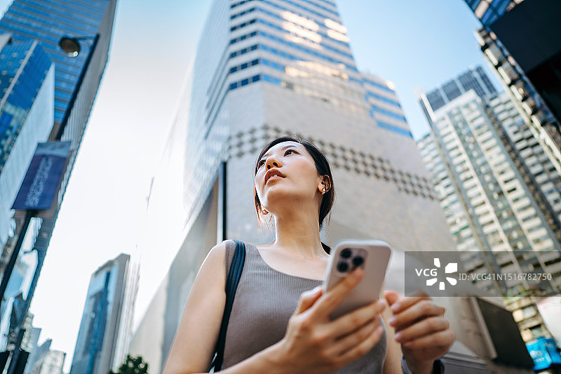 年轻亚洲女商人在市中心街道户外行走时使用智能手机的仰视角度照片，背景是现代摩天大楼和商业广告牌。图片素材