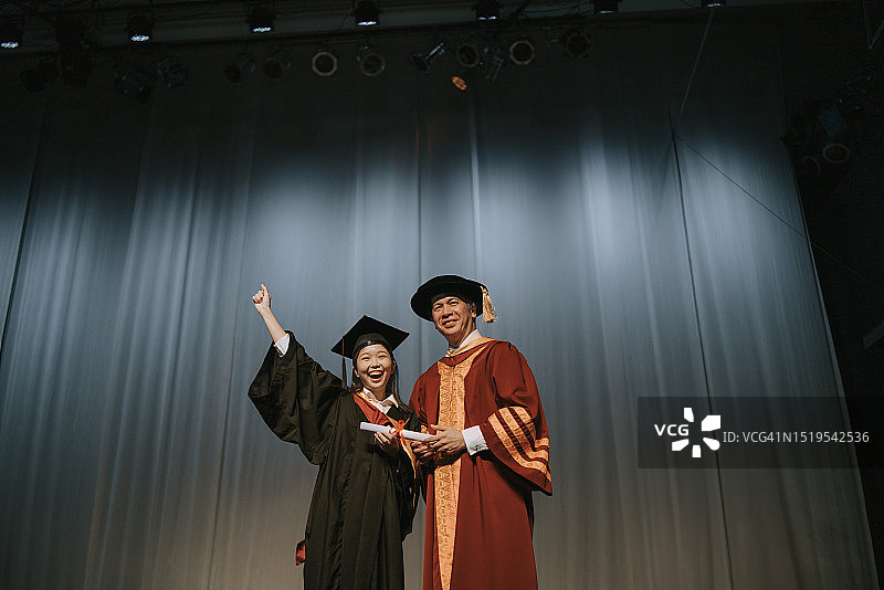 毕业典礼：兴奋的亚洲女大学生在礼堂舞台上收到毕业证书后，站在院长旁边举起手臂图片素材
