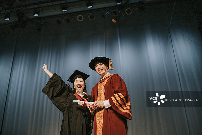 毕业典礼：兴奋的亚洲女大学生在礼堂舞台上收到毕业证书后，站在院长旁边举起手臂图片素材