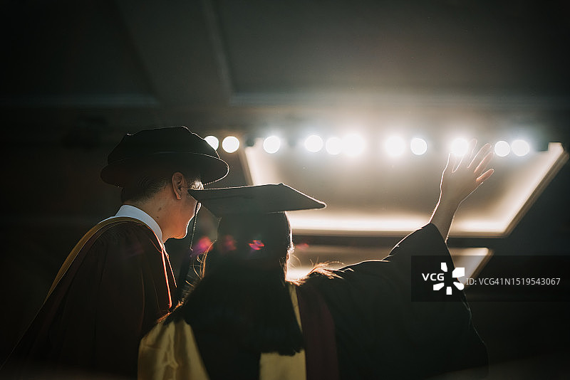 毕业典礼：兴奋的亚洲女大学生在礼堂舞台上收到毕业证书后，站在院长旁边举起手臂图片素材