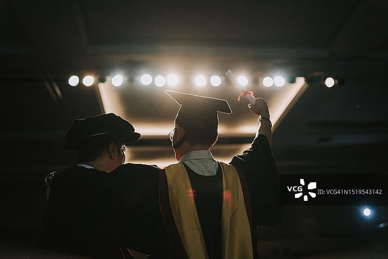 毕业典礼上，兴奋的亚洲毕业生从院长手中接过毕业证书后站在礼堂舞台旁，举起手臂图片素材