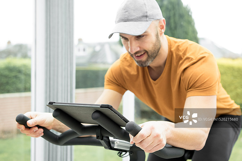 年轻的西班牙裔男子在爱丁堡的阳光房里骑健身车，同时通过平板电脑观看在线健身课程图片素材