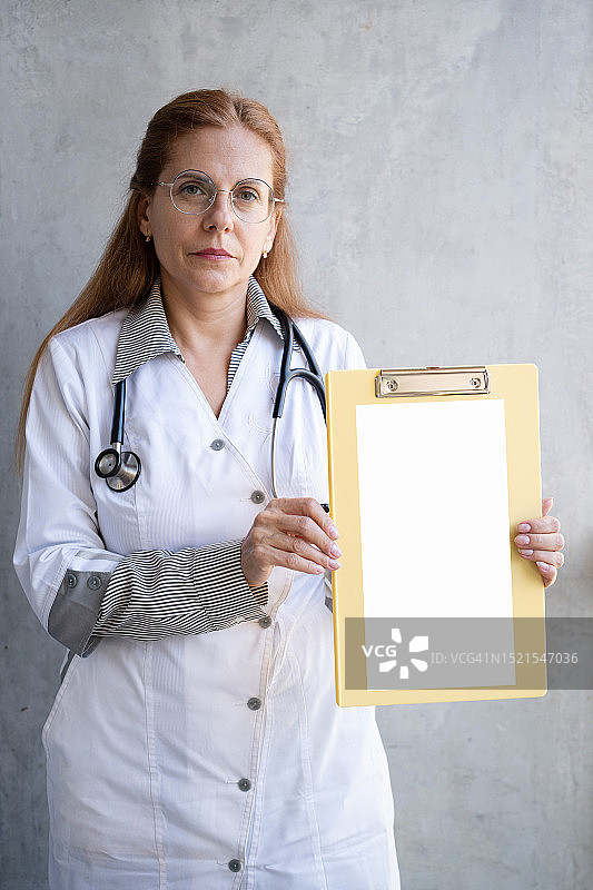 表情严肃、充满自信的女医生拿着写字板靠在灰色墙壁上图片素材