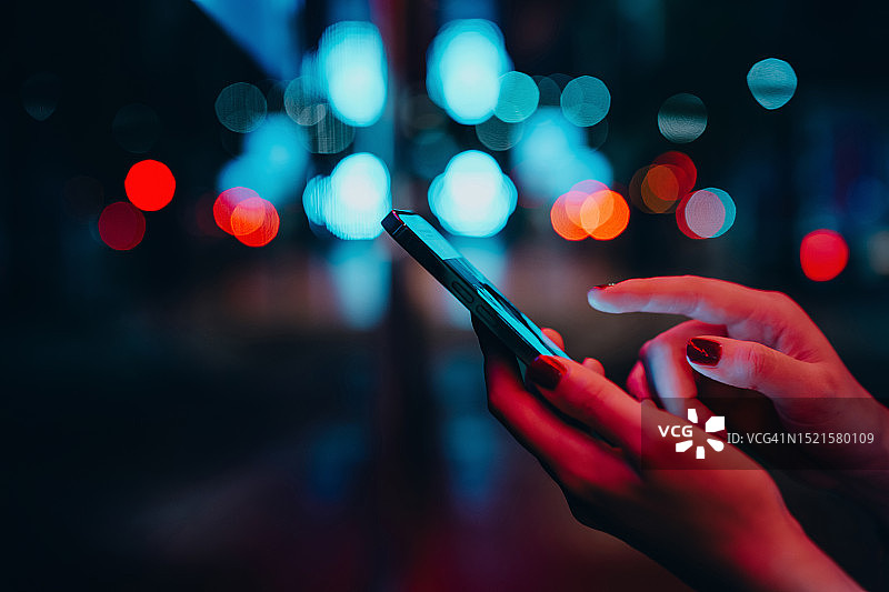 夜晚城市街道上用智能手机进行社交的年轻女子。背景是散焦的街灯。在城市里用智能手机查看社交媒体的女性特写。人们使用科技进行社交。生活方式与科技图片素材