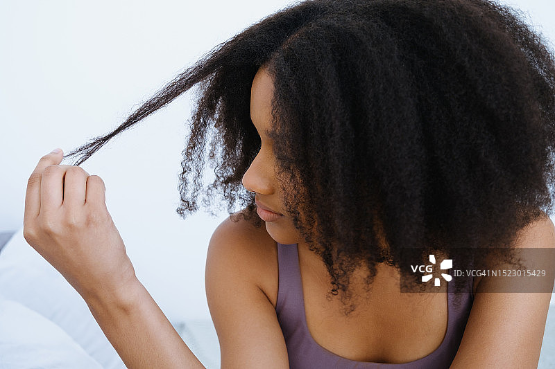 一位美丽的黑人女孩，有着卷曲蓬乱的头发，担心她受损的发梢。一位沮丧的女人用手拿着一缕头发，悲伤地看着它们。头发的问题。沙龙护理或程序不佳。发型毁了。图片素材