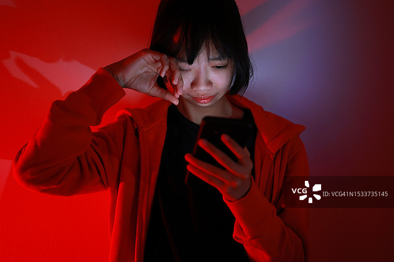一位年轻的亚洲女孩坐在室内地板上，眼泪顺着脸颊流下，手里拿着一部智能手机。这张图片描绘了沉迷和网络欺凌的危险图片素材