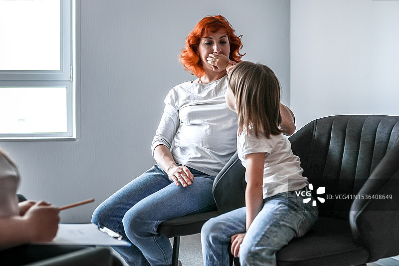 母子沟通困难。母亲和女儿在心理学家诊室，孩子用手捂住母亲的嘴，阻止她说话图片素材