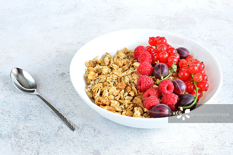 白盘子里放着覆盆子、醋栗和红醋栗的格兰诺拉麦片，配有勺子图片素材