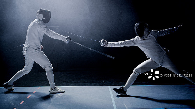 两位职业击剑手在史诗般的蓝色舞台灯光和烟雾效果下，用花剑互相决斗图片素材