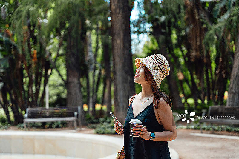 年轻的亚洲女人拿着智能手机和咖啡走在公园里。图片素材