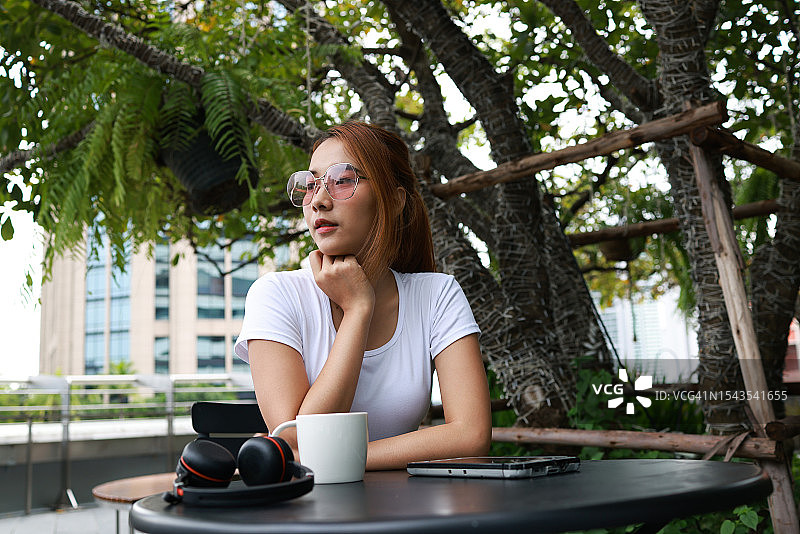 沉浸在思考中的女人：戴着太阳镜，坐在公园的工作场所桌子前，面对平板电脑和耳机，双手托着下巴，望着远方，思考着工作中的点滴。女商人计划或试图解决一些业务图片素材