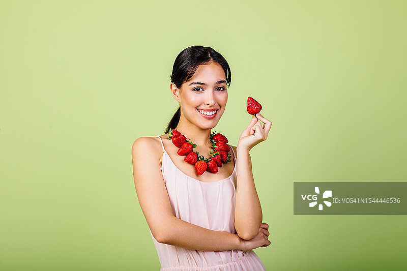 微笑的深色头发女子佩戴草莓配饰，在绿色工作室庆祝自然和谐、身体排毒和健康生活图片素材