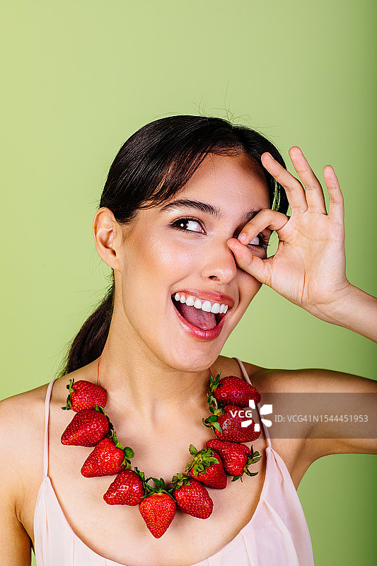 美丽女人在眼睛附近做出OK手势，在绿色工作室里戴着草莓珠子。近距离特写图片素材