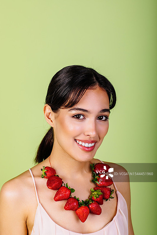拥抱草莓的健康生活方式：微笑的女士戴着草莓项链在绿色背景下看着镜头图片素材