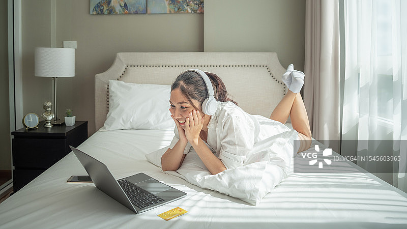 快乐微笑的亚洲女人穿着白色睡衣躺在舒适的床上，用信用卡在笔记本电脑上进行网上购物，早上在卧室里成功支付图片素材