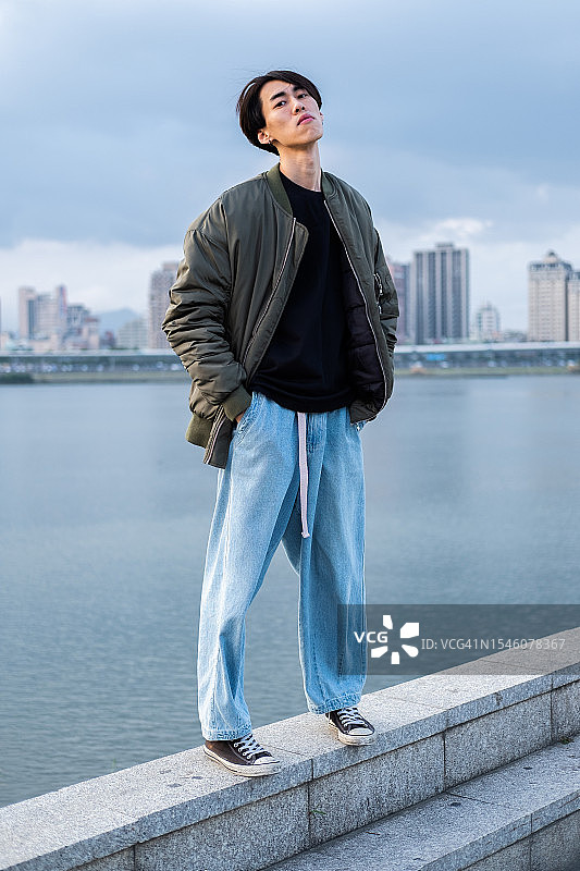 年轻亚洲男子身穿休闲服装，在城市河流附近摆姿势，俯瞰高层建筑。街头风格，都市生活图片素材