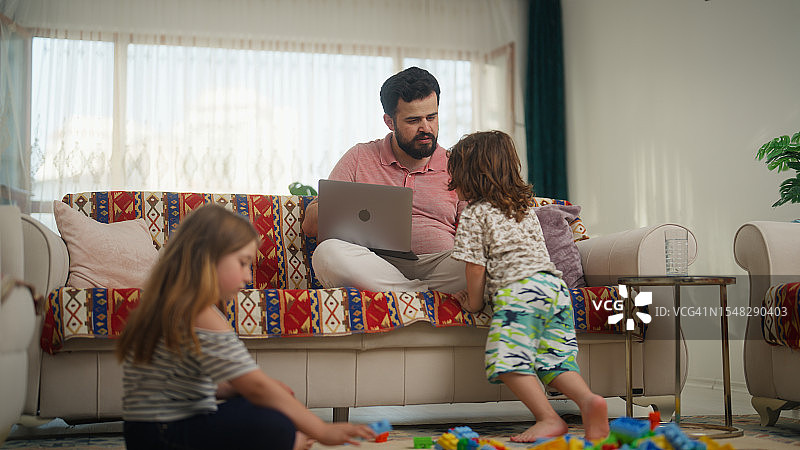 父亲在家用笔记本电脑工作，孩子们在旁边玩耍图片素材