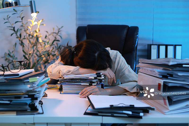 办公室里疲惫的女人趴在桌子上睡着了。大量的文书工作，疲惫的员工，一堆文件和纸张。独自一人昏昏欲睡的女孩在深夜努力工作时在桌子上打瞌睡图片素材