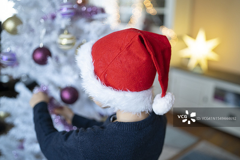 戴着圣诞帽的小男孩将圣诞装饰品放在树上图片素材