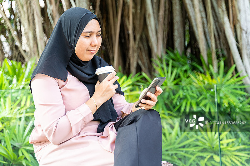 亚洲女人坐在长椅公园里，一边喝咖啡一边看智能手机图片素材