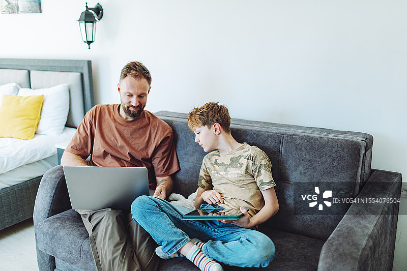 父亲在家用笔记本电脑工作，儿子在旁边使用平板电脑图片素材