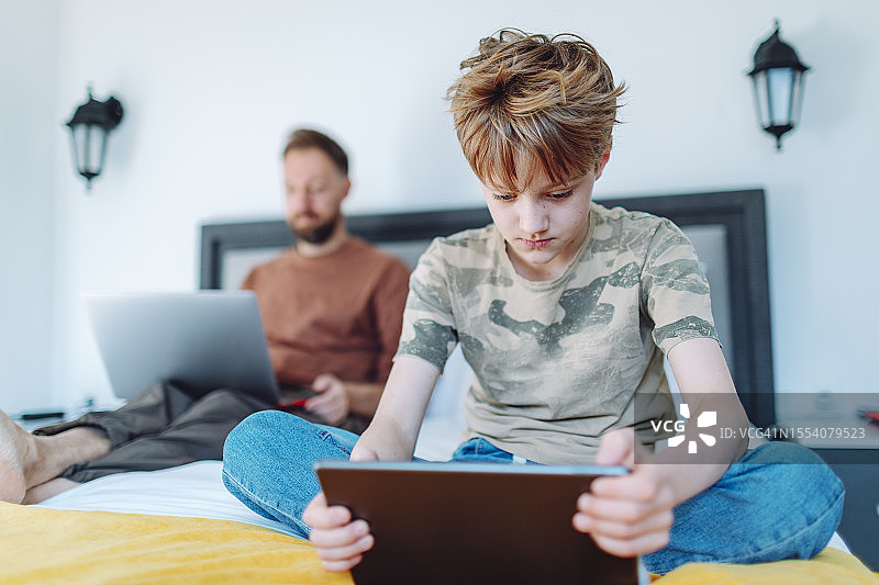 父亲在家用笔记本电脑工作，儿子在旁边使用平板电脑图片素材