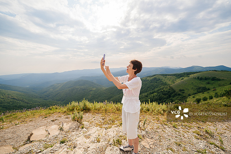 老年妇女在山顶欣赏美丽的大自然，用智能手机与朋友进行视频通话，夏季旅行图片素材