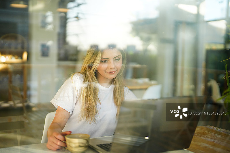 在咖啡馆靠窗的桌旁用笔记本电脑学习的年轻女子图片素材