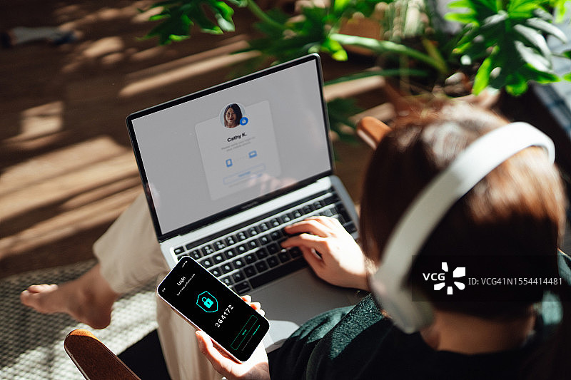 年轻女子通过智能手机和笔记本电脑登录双重身份验证(2FA)在线账户的肩部视角图片素材