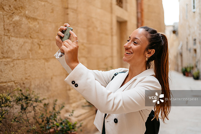 年轻女人在马耳他姆迪纳的巷子里用智能手机拍照图片素材