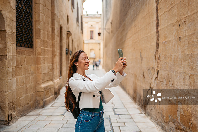 年轻女人在马耳他姆迪纳的巷子里用智能手机拍照图片素材