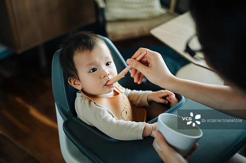 亚洲女婴坐在高脚椅上吃饭，年轻母亲在家喂宝宝图片素材