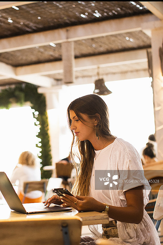 年轻女子在海滩餐厅酒吧的桌旁使用电脑、笔记本电脑和手机工作图片素材