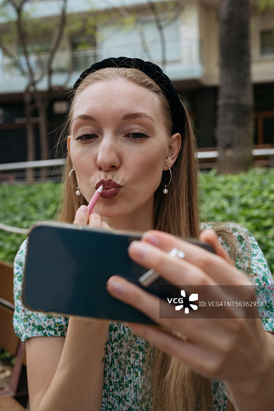 年轻女子在城市街头用智能手机当镜子涂口红图片素材