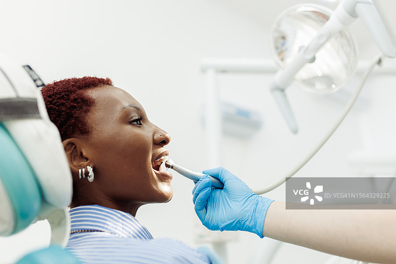 在牙科钻孔过程中，坐在牙科椅上张开嘴的女性患者的侧面照图片素材